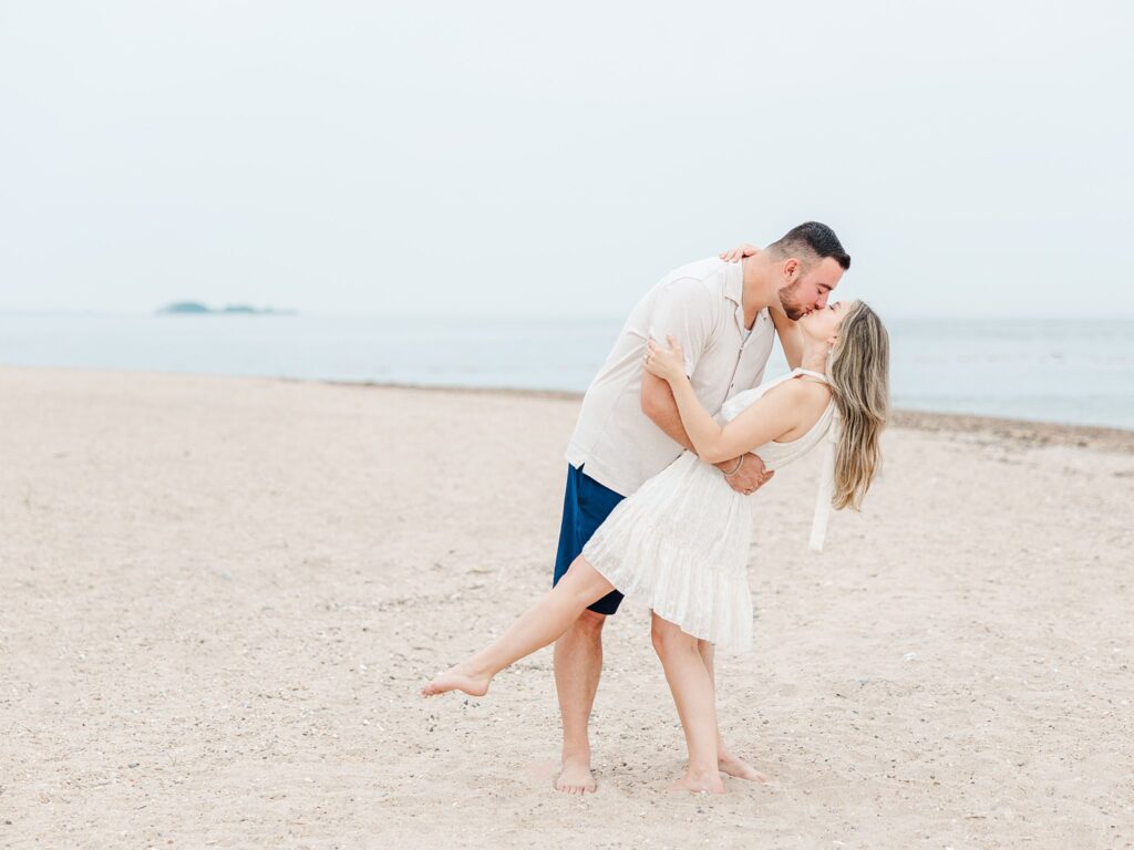 Couple kissing on the beach during a romantic beach engagement session in Clinton Connecticut under cloudy skies